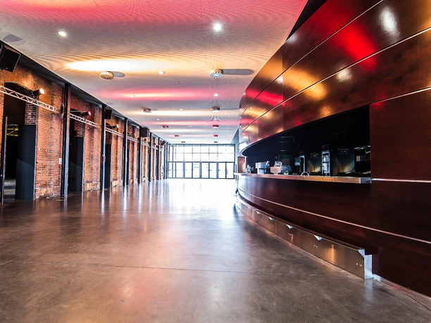 Palladium Köln Foyer Das Bild zeigt das Foyer des Palladium Köln. Der Raum hat eine geschwungene Holztheke rechts und offen gelegte Backsteinwände links. Die Decke ist mit einer modernen Lichtinstallation beleuchtet, die warme, rötliche Töne widergibt. Der Boden besteht aus glattem Beton, was eine moderne, industrielle Atmosphäre schafft. Im Hintergrund befindet sich ein großer Fensterbereich, durch den Tageslicht einfällt und den Raum erhellt.