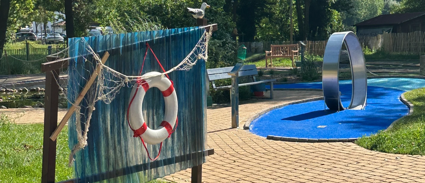 Adventuregolf am Meer Ein kurviger, gepflasterter Weg führt zu einer modernen Adventuregolfanlage, umgeben von Bäumen.