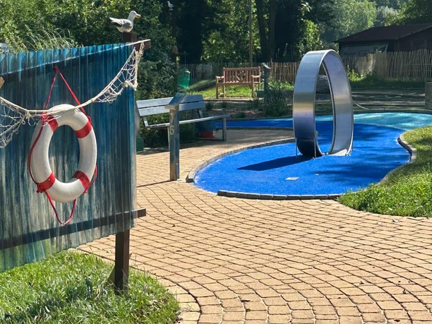 Adventuregolf am Meer Ein kurviger, gepflasterter Weg führt zu einer modernen Adventuregolfanlage, umgeben von Bäumen.