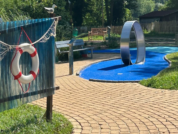 Adventuregolf am Meer Ein kurviger, gepflasterter Weg führt zu einer modernen Adventuregolfanlage, umgeben von Bäumen.