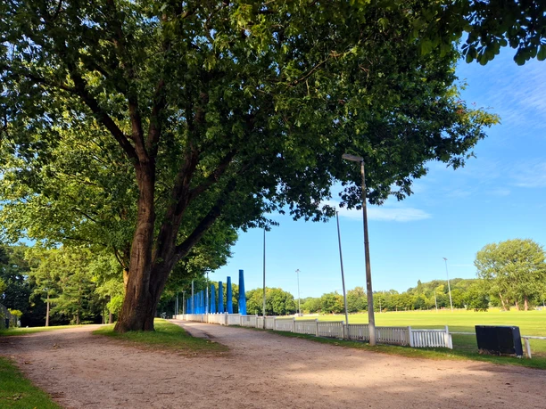 Turnierplatz Tribuenen.jpg Das Bild zeigt eine breite, von Bäumen gesäumte Allee neben einem offenen Sportplatz mit blau-weißen Tribünen.