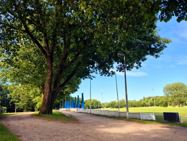 Turnierplatz Tribuenen.jpg Das Bild zeigt eine breite, von Bäumen gesäumte Allee neben einem offenen Sportplatz mit blau-weißen Tribünen.