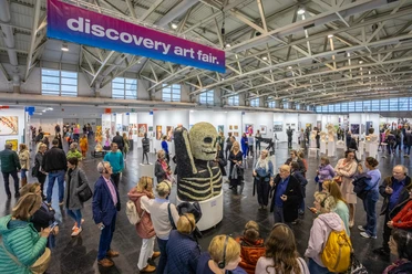 Discovery Art Fair in Frankfurt am Main mit Besucher*innen und Kunstwerken Menschen auf der Discovery Art Fair in der Messe Frankfurt betrachten Kunstwerke, darunter eine große Skelett-Skulptur, in einer hellen, weitläufigen Halle.