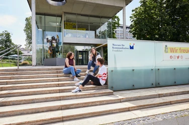 Drei Jugendliche sitzen entspannt auf den Treppen vor dem modernen Eingangsgebäude des Museums für Kommunikation Frankfurt. Three young people sitting relaxed on the stairs in front of the modern entrance building of the Museum of Communication Frankfurt.