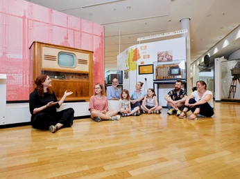 Gruppenführung im Museum für Kommunikation Frankfurt vor historischem Fernseher Eine Gruppe von Besucher*innen sitzt auf dem Boden vor einem historischen Fernseher und hört aufmerksam einer Führerin zu.A group of visitors sitting on the floor in front of a historic TV, attentively listening to a guide.