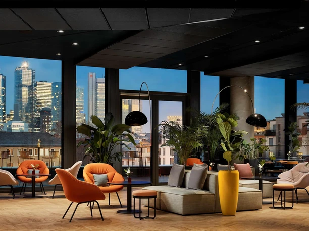 OAKS Bar im NH Collection Frankfurt Spin Lounge mit orangenen Sesseln, Pflanzen und Blick auf beleuchtete Skyline.