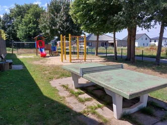 Das Bild zeigt einen kleinen Spielplatz mit einer Rutsche, einer Schaukel und einem alten Tischtennistisch.