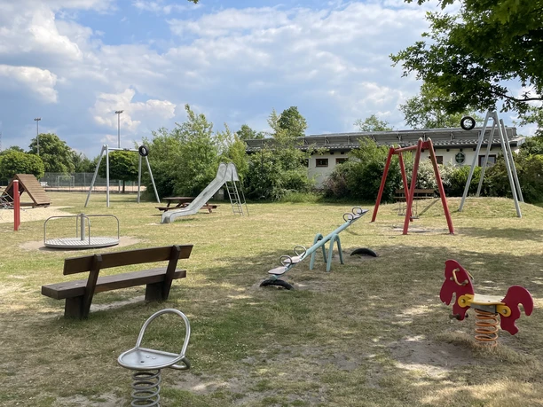 Grüne Wiese mit Spielgeräten: Schaukeln, Rutschen und Wippen unter Bäumen und blauem Himmel.