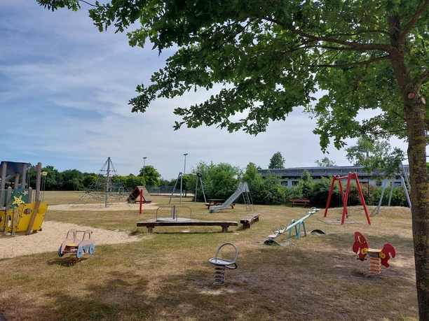 Der Spielplatz mit Rutschen, Schaukeln und Wippen lädt zum Spielen ein. Schatten spendet ein Baum.