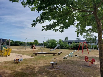 Begegnungsstätte Wehe Der Spielplatz mit Rutschen, Schaukeln und Wippen lädt zum Spielen ein. Schatten spendet ein Baum.