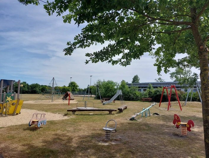 Begegnungsstätte Wehe Der Spielplatz mit Rutschen, Schaukeln und Wippen lädt zum Spielen ein. Schatten spendet ein Baum.
