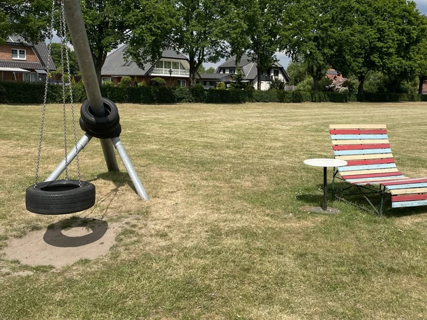 Begegnungsstätte Wehe Im Bild ist eine große Wiese zu sehen, auf der rechts eine rot-blaue Bank mit kleinem Tisch steht. Links im Vordergrund befindet sich eine metallene Spielgeräte-Aufhängung mit einem Autoreifen als Schaukel. Im Hintergrund sind Bäume und einige Häuser erkennbar.