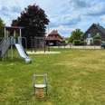 Spielplatz mit Rutsche und Schaukeltier auf grüner Wiese, umgeben von Bäumen und Häusern.