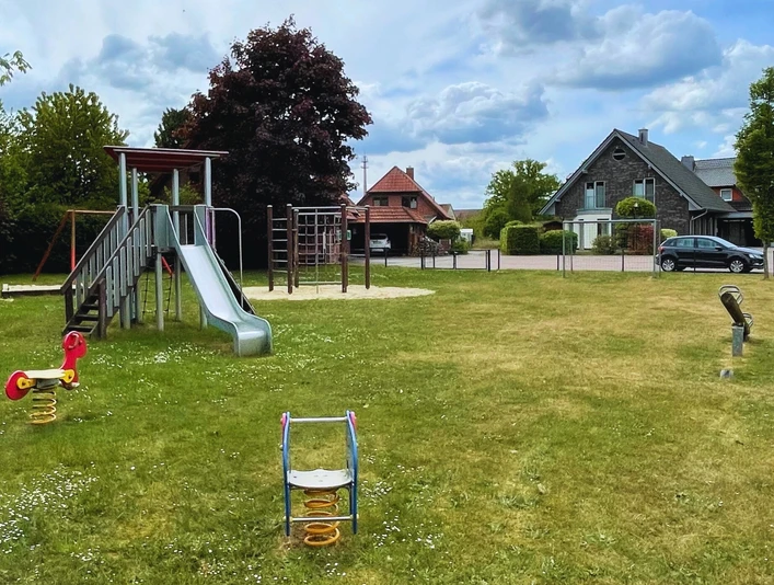 Spielplatz mit Rutsche und Schaukeltier auf grüner Wiese, umgeben von Bäumen und Häusern.