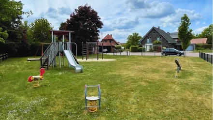 Mühlenweg Spielplatz mit Rutsche und Schaukeltier auf grüner Wiese, umgeben von Bäumen und Häusern.