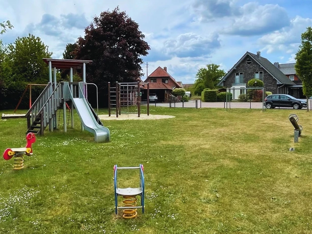 Mühlenweg Spielplatz mit Rutsche und Schaukeltier auf grüner Wiese, umgeben von Bäumen und Häusern.