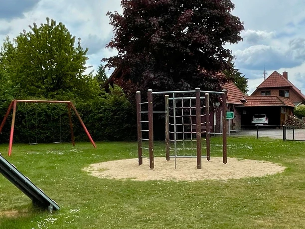 Mühlenweg Ein Spielplatz mit Rutsche, Klettergerüst und Schaukel auf einer Wiese. Hintergrund: Häuser.