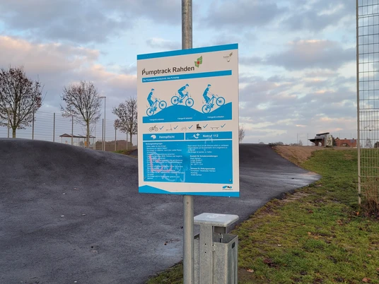 Das Bild zeigt einen Pumptrack mit Hinweisschild in Rahden. Die Strecke aus Asphalt windet sich über hügeliges Gelände.