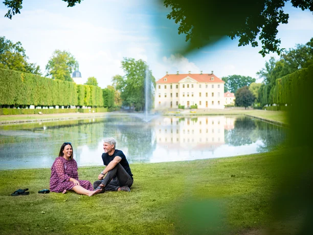 ZAB_AndreForner_Teich_7664_2022.jpg Ein Paar sitzt entspannt am Ufer eines Teichs mit einem historischen Gebäude im Hintergrund.
