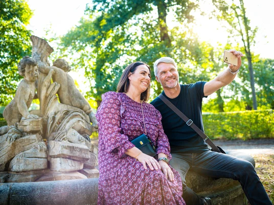 ZAB_AndreForner_Rondell_Flora_7828_2022.jpg Ein Paar sitzt auf einer steinernen Bank im Park, posiert für ein Selfie vor einer Skulptur.A couple sits on a stone bench in the park, posing for a selfie in front of a sculpture.Pár sedí na kamenné lavičce v parku a pózuje pro selfie před sochou.Para siedzi na kamiennej ławce w parku, pozując do selfie przed rzeźbą.Een koppel zit op een stenen bankje in het park en poseert voor een selfie voor een beeldhouwwerk.Una coppia si siede su una panchina di pietra nel parco, in posa per un selfie davanti a una scultura.