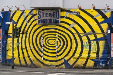 Stereo Wonderland The picture shows the front of a building with a striking yellow and blue spiral pattern. There is a sign on the façade that reads "Stereo Wonderland". Graffiti and posters can be seen on the roller shutter and the walls, creating an urban and artistic atmosphere.