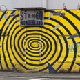 Stereo Wonderland Das Bild zeigt die Vorderseite eines Gebäudes mit einem markanten gelben und blauen Spiralmuster. An der Fassade befindet sich ein Schild mit der Aufschrift "Stereo Wonderland". Graffiti und Plakate sind auf dem Rolltor und den Wänden zu sehen, was eine urbane und künstlerische Atmosphäre erzeugt.The picture shows the front of a building with a striking yellow and blue spiral pattern. There is a sign on the façade that reads "Stereo Wonderland". Graffiti and posters can be seen on the roller shutter and the walls, creating an urban and artistic atmosphere.