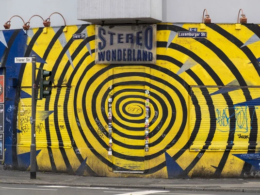 Stereo Wonderland Das Bild zeigt die Vorderseite eines Gebäudes mit einem markanten gelben und blauen Spiralmuster. An der Fassade befindet sich ein Schild mit der Aufschrift "Stereo Wonderland". Graffiti und Plakate sind auf dem Rolltor und den Wänden zu sehen, was eine urbane und künstlerische Atmosphäre erzeugt.The picture shows the front of a building with a striking yellow and blue spiral pattern. There is a sign on the façade that reads "Stereo Wonderland". Graffiti and posters can be seen on the roller shutter and the walls, creating an urban and artistic atmosphere.