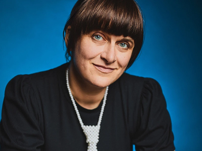 Ella_Carina_Werner_Foto_Julia_Schwendner.jpg Portrait einer Frau mit braunen Haaren und blauem Hintergrund, die eine Perlenkette trägt.