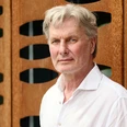 NorbertHorst_2025_JoachimGrothus.jpg Ein Mann mit grauen Haaren und weißem Hemd steht vor einer modernen Wand mit wellenförmigen Öffnungen.