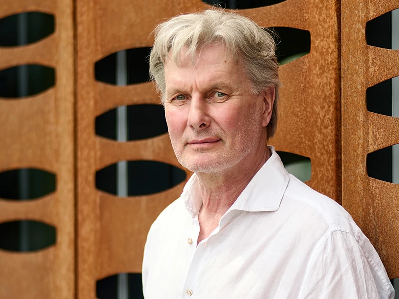 NorbertHorst_2025_JoachimGrothus.jpg Ein Mann mit grauen Haaren und weißem Hemd steht vor einer modernen Wand mit wellenförmigen Öffnungen.