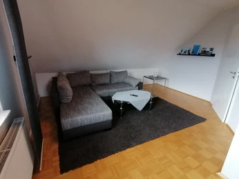 IMG_20250724_153219.jpg Wohnzimmer mit grauem Ecksofa, kleinem Glastisch und Teppich auf hellem Holzboden unter Dachschräge.