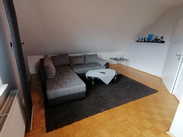 IMG_20250724_153219.jpg Wohnzimmer mit grauem Ecksofa, kleinem Glastisch und Teppich auf hellem Holzboden unter Dachschräge.