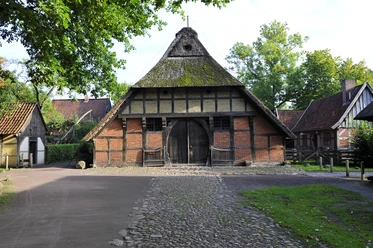 Fachwerkbau mit Strohdach und großer Holztür vor gepflastertem Weg, umgeben von Bäumen.