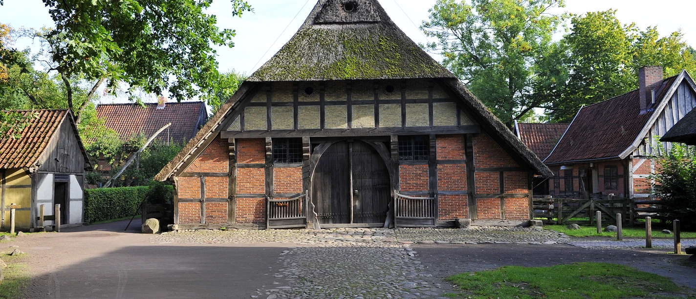 Fachwerkbau mit Strohdach und großer Holztür vor gepflastertem Weg, umgeben von Bäumen.