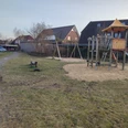 Spielplatz an den Weiden Ein Spielplatz An den Weiden mit Holz-Klettergerüst und Rutsche, umgeben von Gras und einigen Häusern im Hintergrund.