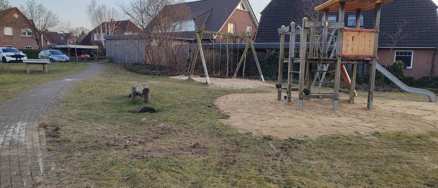 Spielplatz an den Weiden Ein Spielplatz An den Weiden mit Holz-Klettergerüst und Rutsche, umgeben von Gras und einigen Häusern im Hintergrund.