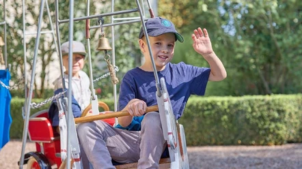 Museumsfest Glashütte Gernheim.jpg Ein lächelnder Junge im Spielgerät grüßt, während im Hintergrund ein älterer Herr sitzt.