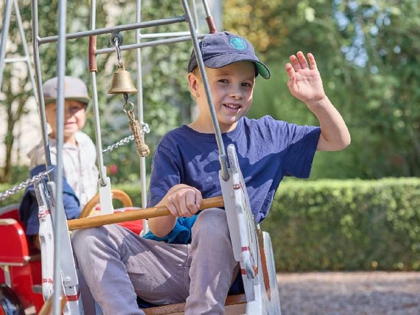 Museumsfest Glashütte Gernheim.jpg Ein lächelnder Junge im Spielgerät grüßt, während im Hintergrund ein älterer Herr sitzt.