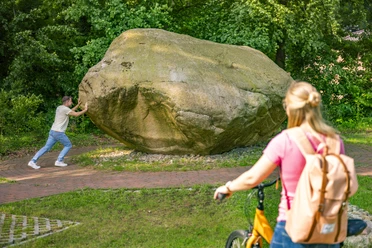 Koloss von Hüven – Radfahren Hümmling ©Emsland Tourismus GmbH (2).jpg Großer Findling im Grünen, ein Mann stützt sich dagegen, Frau mit Fahrrad betrachtet die Szene.