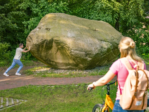 Koloss von Hüven – Radfahren Hümmling ©Emsland Tourismus GmbH (2).jpg Großer Findling im Grünen, ein Mann stützt sich dagegen, Frau mit Fahrrad betrachtet die Szene.