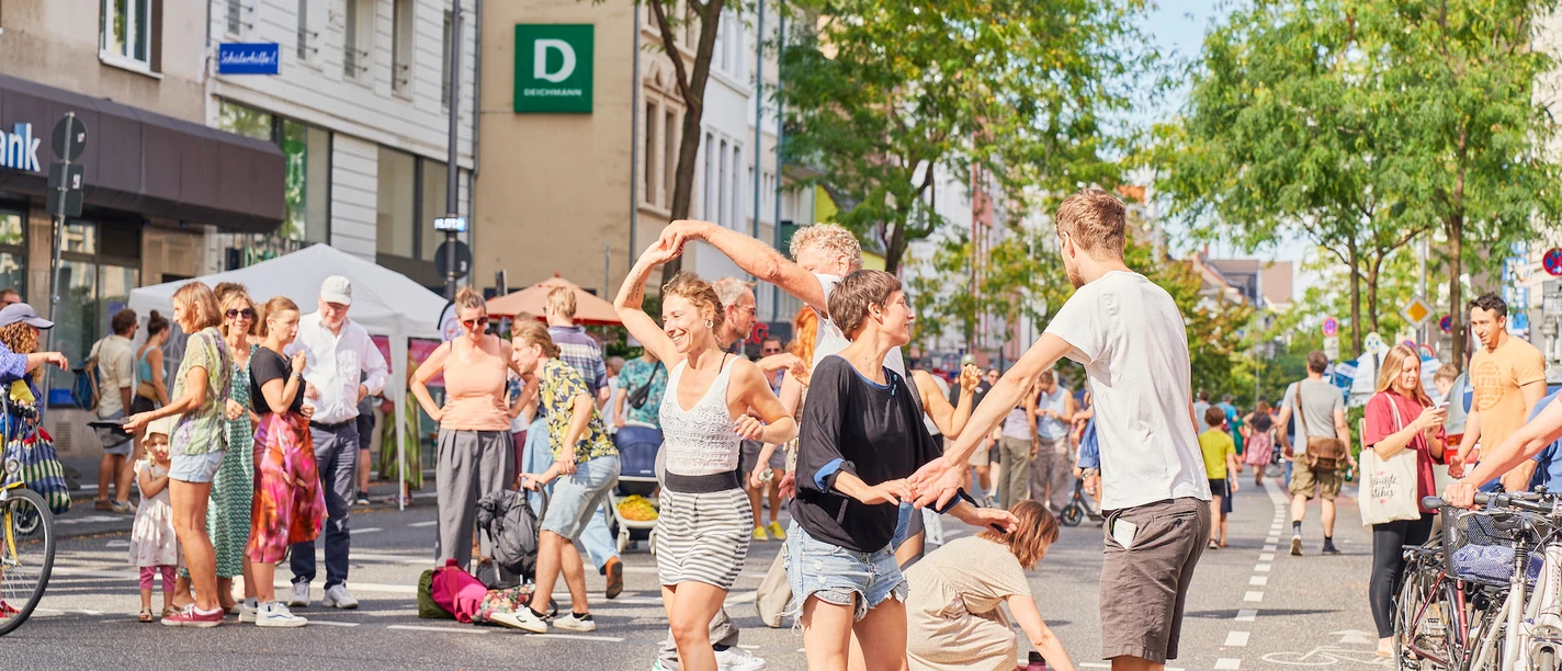 Day of the good life Street party with people dancing and celebrating together, surrounded by trees and buildings.