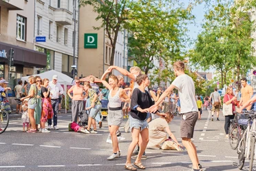 Tag des guten Lebens Straßenfest mit Menschen, die zusammen tanzen und fröhlich feiern, umgeben von Bäumen und Gebäuden.