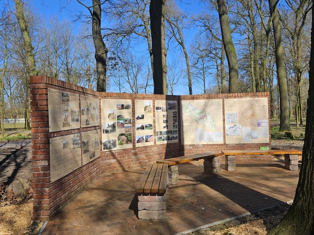 Wehmer Geschichtspfad, Werlte ©Emsland Tourismus GmbH (17).jpg Informationstafeln mit historischen Bildern und Texten auf einer Ziegelwand im Wald bei Sonnenschein