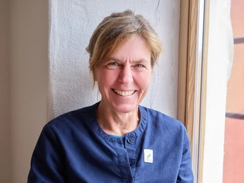 Zugeschnitten_nain_design.jpg Eine lächelnde Person mit blauer Kleidung sitzt am Fenster.
