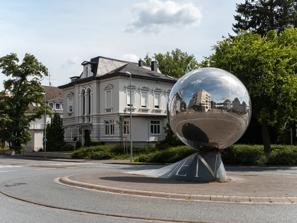 Kunstwerk aus Edelstahl, reflektiert umliegende Gebäude und Himmel, platziert in einem Kreisverkehr.