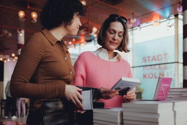 lit.COLOGNE Zwei Frauen studieren Bücher in einem modernen Ausstellungsraum. Im Hintergrund leuchtet "Stadthalle Köln".
