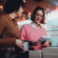 lit.COLOGNE Zwei Frauen studieren Bücher in einem modernen Ausstellungsraum. Im Hintergrund leuchtet "Stadthalle Köln".Two women study books in a modern exhibition space. "Stadthalle Köln" shines in the background.
