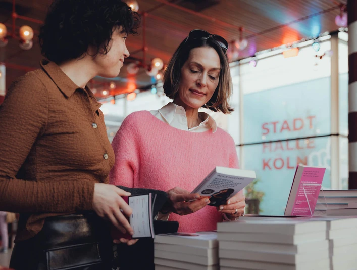 lit.COLOGNE Zwei Frauen studieren Bücher in einem modernen Ausstellungsraum. Im Hintergrund leuchtet "Stadthalle Köln".Two women study books in a modern exhibition space. "Stadthalle Köln" shines in the background.