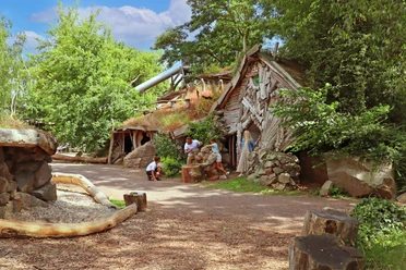 Children explore a grass-covered entrance to an abandoned wooden mine, surrounded by trees.