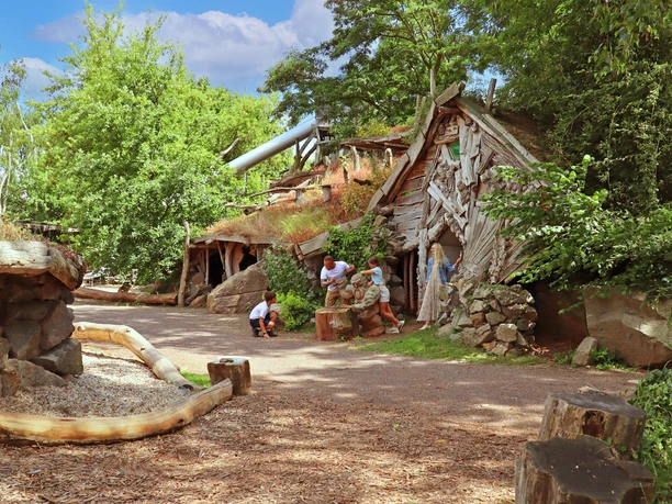 Children explore a grass-covered entrance to an abandoned wooden mine, surrounded by trees.